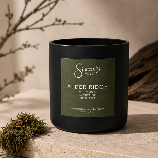 A black candle jar with a green label reading 'Sincerely, Man Alder Ridge' on a stone surface, styled with dried branches and moss in a warm neutral setting.