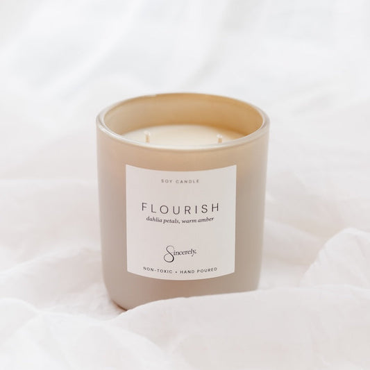 A soy candle in a matte taupe-beige glass jar with a white label reading 'FLOURISH', set against a soft white draped fabric background.