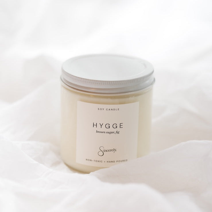 A soy candle in a clear glass jar with a white lid and a minimalist white label reading 'HYGGE' set against a soft white draped fabric background.