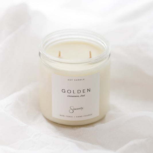 A soy candle in a clear glass jar with a cream-colored wax and a white label reading 'GOLDEN' and 'Sincerely', set against a soft white draped fabric background.