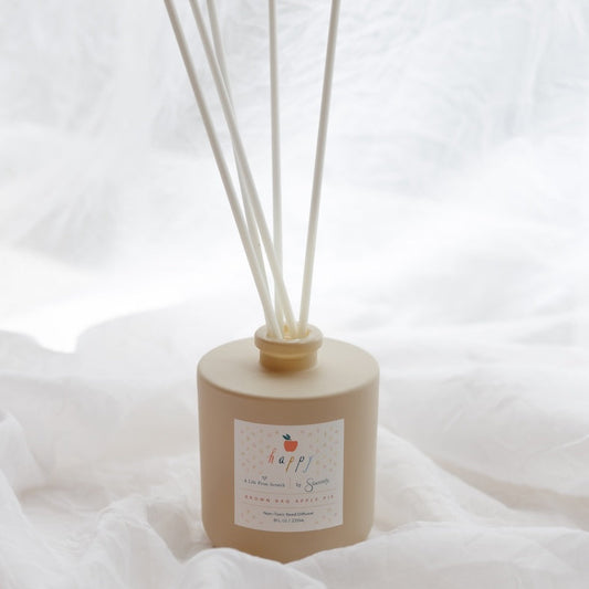 A reed diffuser bottle in a matte beige finish with natural reed sticks, resting on a soft white fabric background.