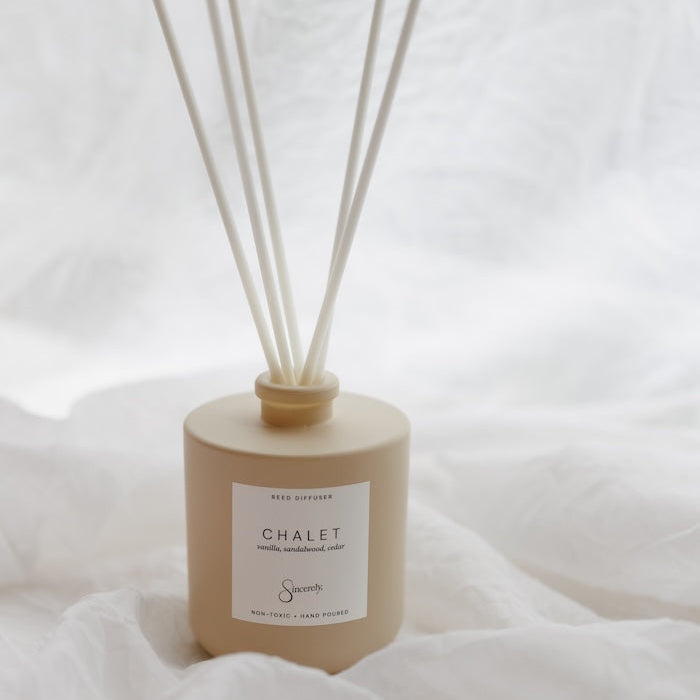 A beige reed diffuser bottle with six natural reed sticks, labeled CHALET, set against a soft white draped fabric background.