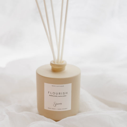 A beige reed diffuser bottle with six natural reed sticks, resting on white draped fabric. The bottle features a white label with the scent 'FLOURISH'.