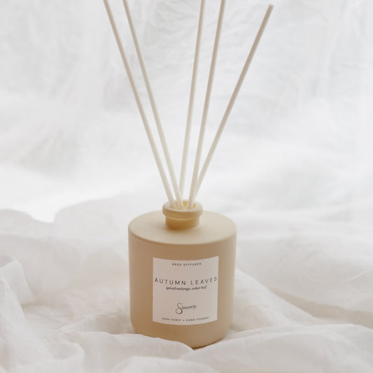 A reed diffuser bottle in a matte beige finish with six natural reed sticks, set against a soft white draped fabric background. The label reads 'AUTUMN LEAVES'