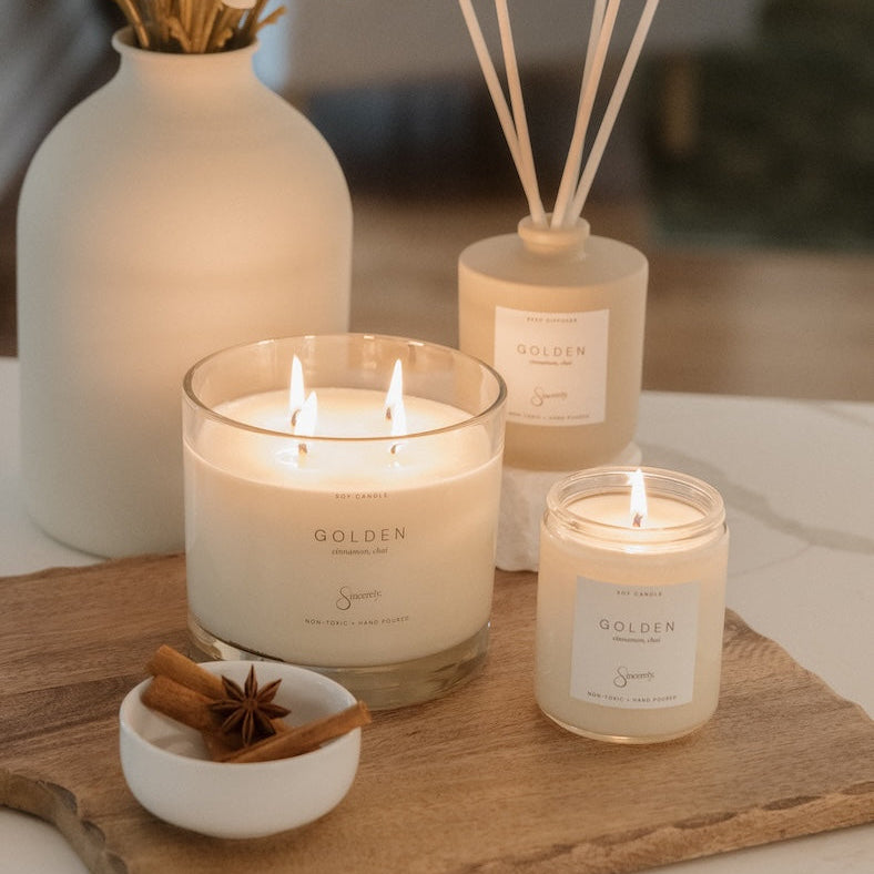 A styled home scene featuring a set of Golden scented home fragrance products, including a large 4-wick candle, a smaller jar candle, and a reed diffuser, arranged on a wooden cutting board next to a white vase with dried flowers.