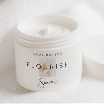 A close-up of a person applying white body butter cream to their hand, with a white jar of 'Flourish' body butter by 'Sincerely' resting on a soft white towel nearby.