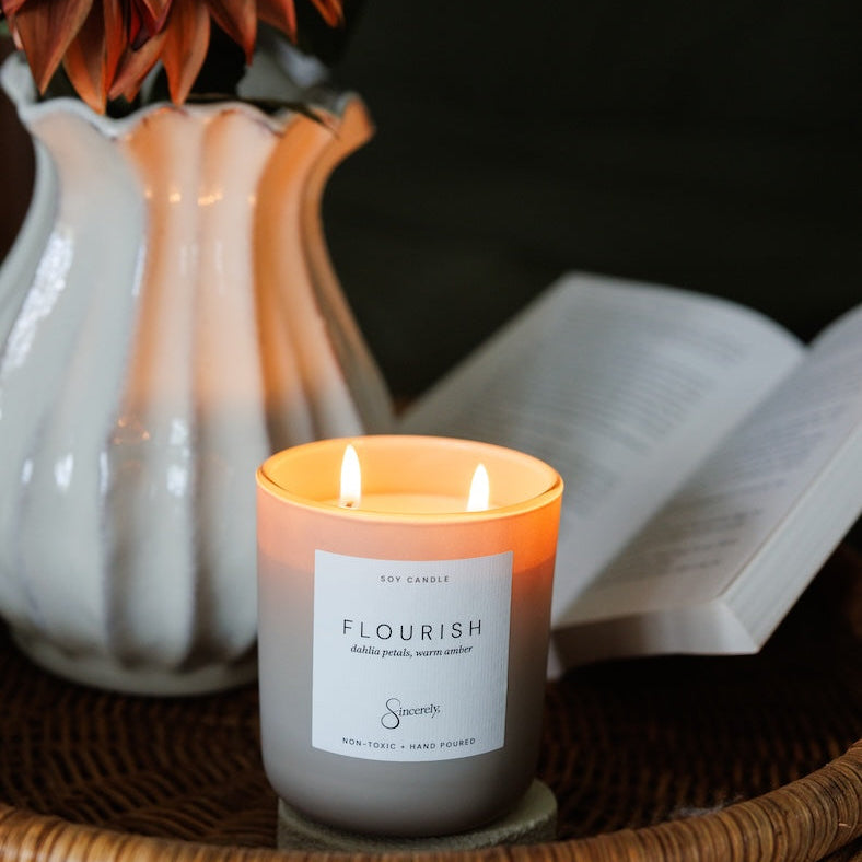 A lit soy candle in a frosted glass jar sits on a woven tray, with a white vase of flowers and an open book softly blurred in the background.
