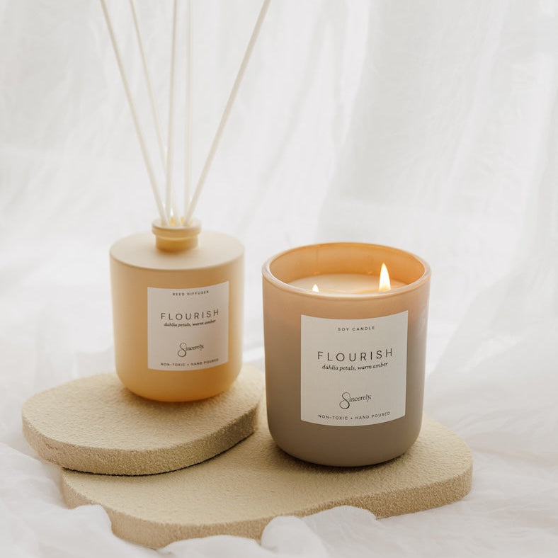 A reed diffuser and a lit scented candle both in warm beige and peachy-orange tones with white labels, styled on a textured beige stone tray against a draped white fabric background.