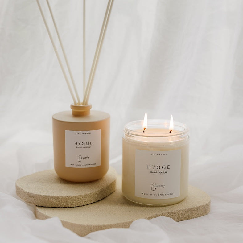 a beige reed diffuser bottle with reeds and a matching double-wick candle, both with cream labels reading 'HYGGE' displayed on round textured stone risers against a draped white fabric background.