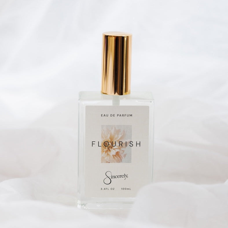 A clear glass perfume bottle with a shiny gold cylindrical cap, labeled 'FLOURISH' by the brand 'Sincerely', set against a soft, draped white fabric background.