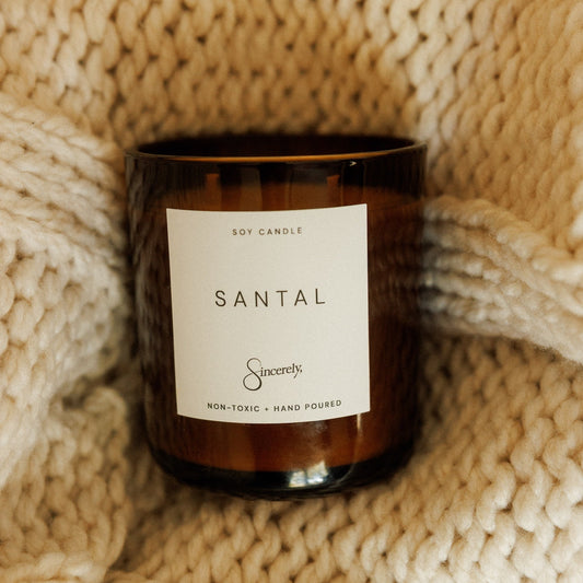 An amber glass soy candle with a white label reading 'SANTAL' and the brand 'Sincerely', styled on a chunky cream knit blanket.
