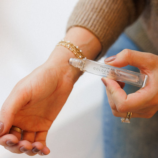 A close-up lifestyle shot of a person holding a clear glass roller bottle labeled 'GOLDEN HOUR PM' over their wrist, showing the clear liquid inside. The person is wearing a beige ribbed sweater, a gold beaded bracelet, and rings.