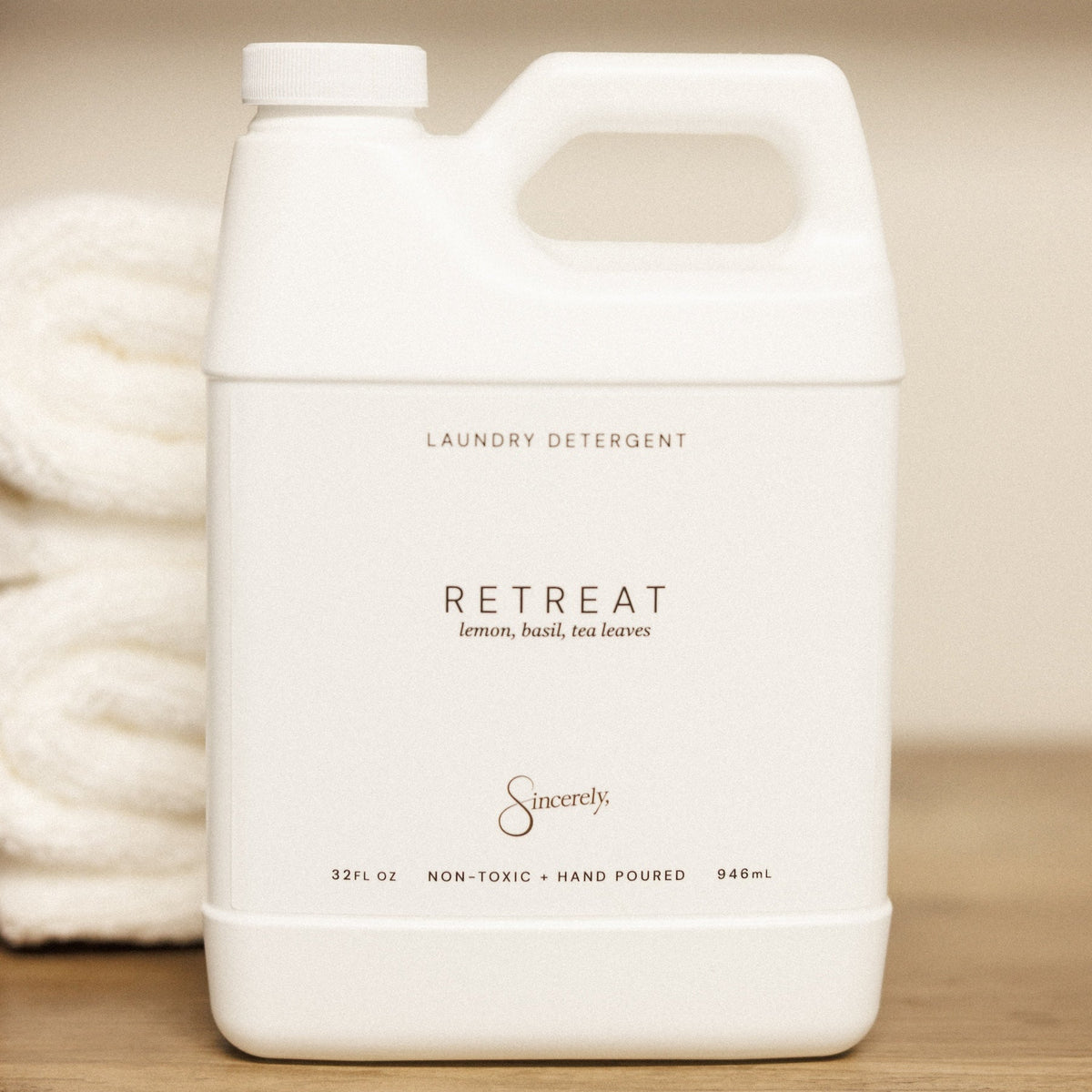 A white plastic jug of Retreat laundry detergent by Sincerely sits on a wooden surface next to a folded white towel, with a soft beige background.