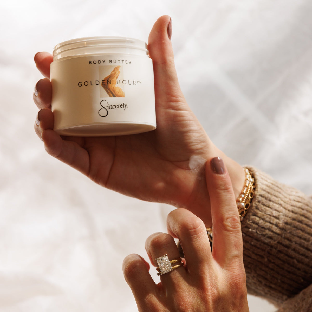 A person holds a cream-colored jar of Gold Hour Body Butter against a soft white fabric background, with their hand near the jar and wearing a beige knit sweater and gold jewelry.