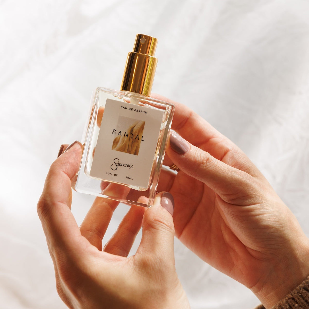A hand holding a clear glass perfume bottle with a gold spray top and a cream label that reads 'SANTAL' and 'Sincerely'. The background is a soft, draped white fabric.