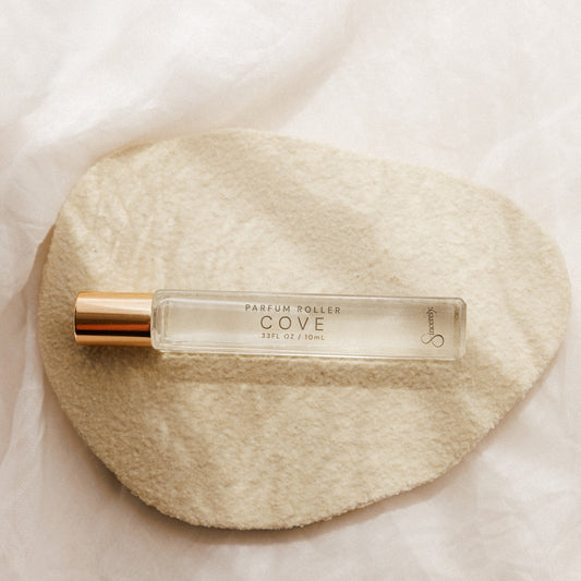 A clear glass perfume rollerball bottle with a metallic gold cap, labeled 'COVE', resting on a textured beige fabric pad over a soft white background.