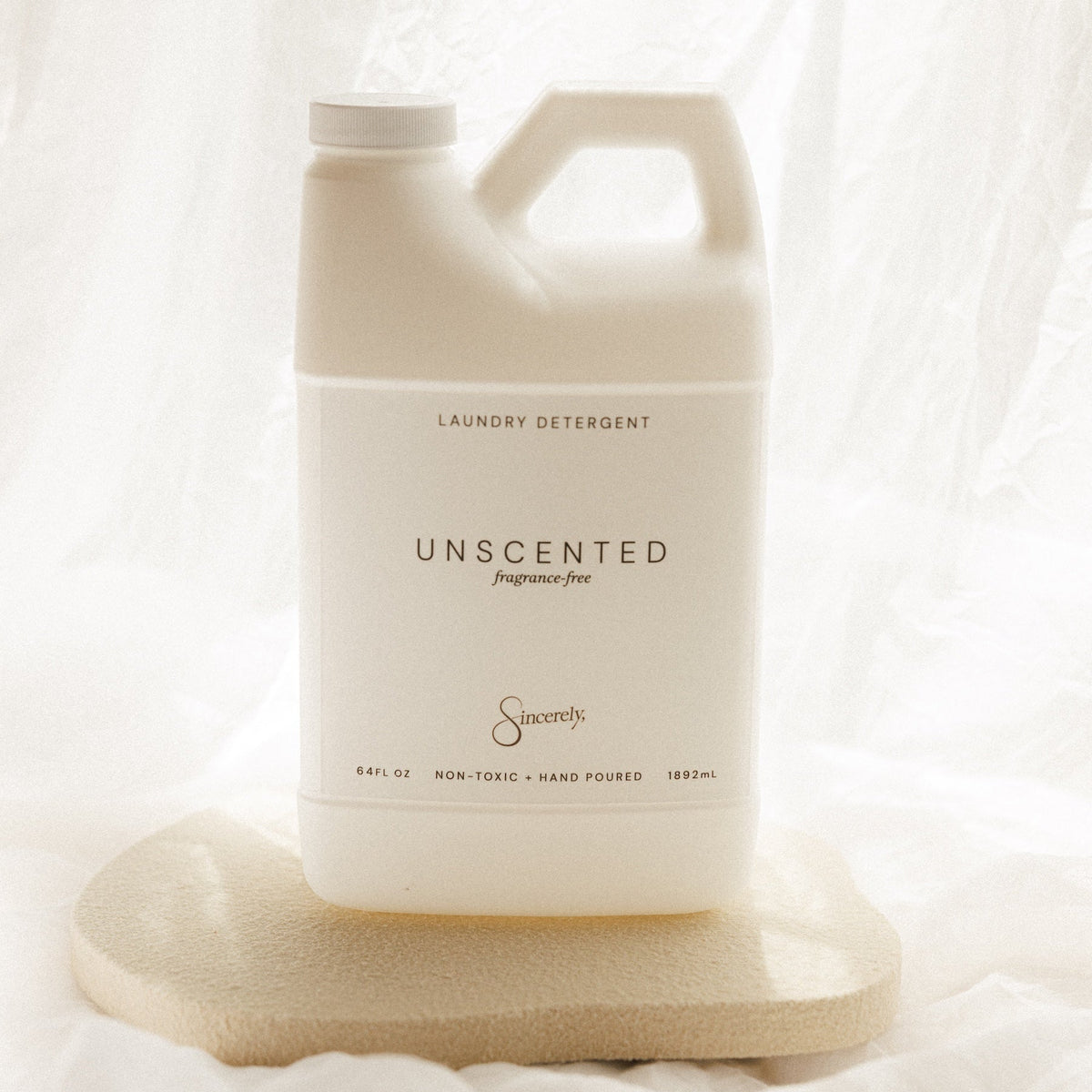 A large white plastic jug of Laundry Detergent, Unscented, by the brand Sincerely, displayed on a textured stone coaster against a draped white fabric background.