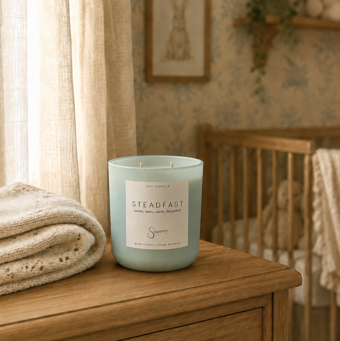 A blue candle in a frosted glass jar sits on a wooden dresser in a softly lit nursery. The front label reads 'STEADFAST', and the room includes patterned wallpaper, a crib, curtains, and folded knit blankets.
