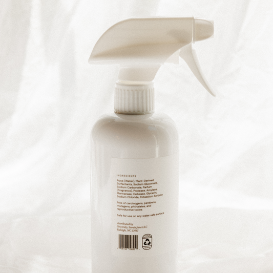 A white plastic spray bottle with a white trigger sprayer, photographed from the back against a soft, draped white fabric background.