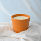 An orange terracotta candle holder containing cream-colored wax with two wicks, styled on draped white fabric against a soft light blue background.