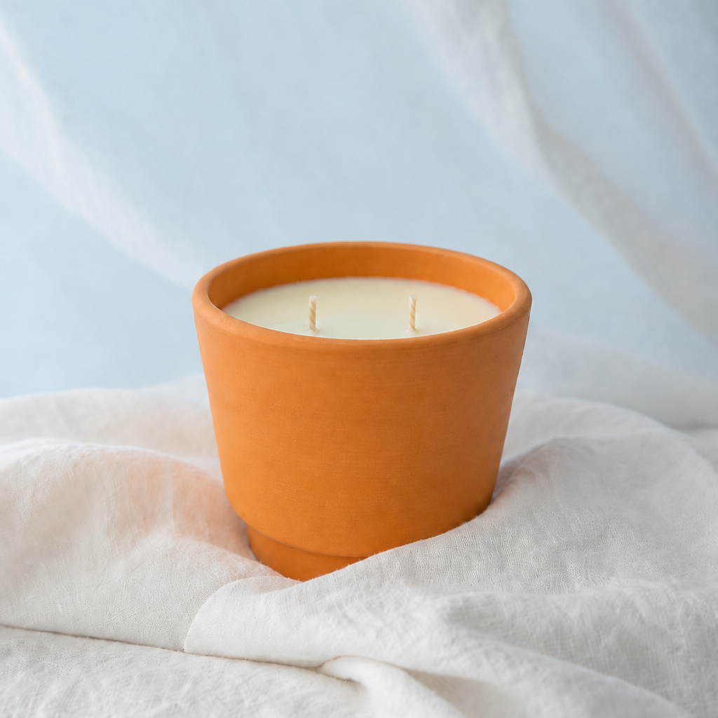 An orange terracotta candle holder containing cream-colored wax with two wicks, styled on draped white fabric against a soft light blue background.