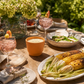 An outdoor dining table styled with a terracotta candle, pink cocktails with peach garnish, a platter of grilled corn, salads, and a large vase of hydrangeas in a lush garden setting.