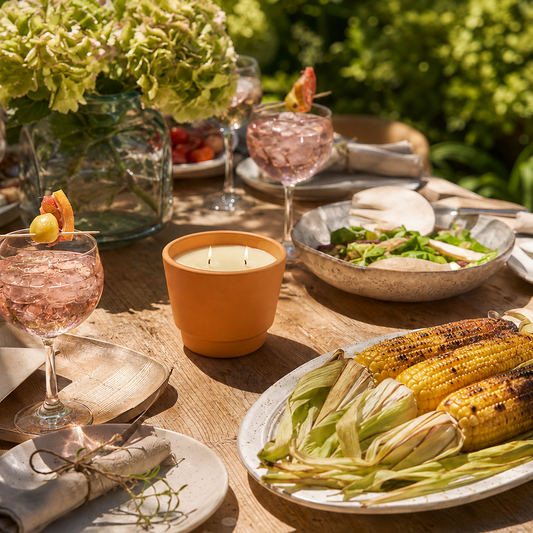 An outdoor dining table styled with a terracotta candle, pink cocktails with peach garnish, a platter of grilled corn, salads, and a large vase of hydrangeas in a lush garden setting.