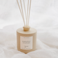 A beige reed diffuser bottle with five natural reed sticks, resting on a soft white fabric background. The bottle features a white label with the text 'HARVEST'.