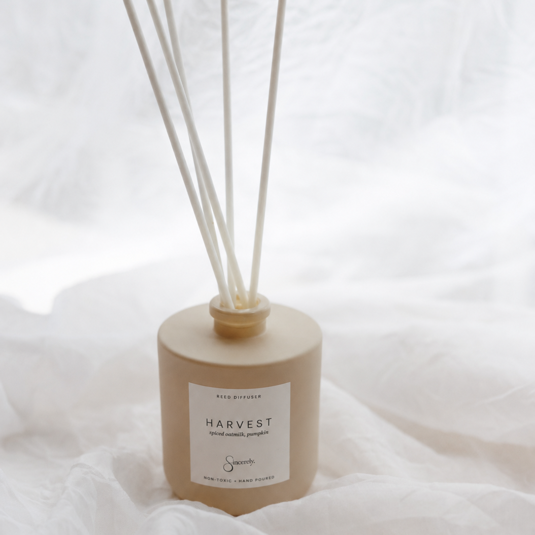 A reed diffuser bottle in a matte beige finish with six natural reed sticks, resting on a soft, draped white fabric background. The label reads 'HARVEST' 
