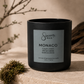A matte black candle jar with a silver label reading 'Sincerely, MAN' and 'MONACO Clean Citrus Spiced Amber Sensual Musk', styled on a stone surface with moss and branches in a warm neutral setting.