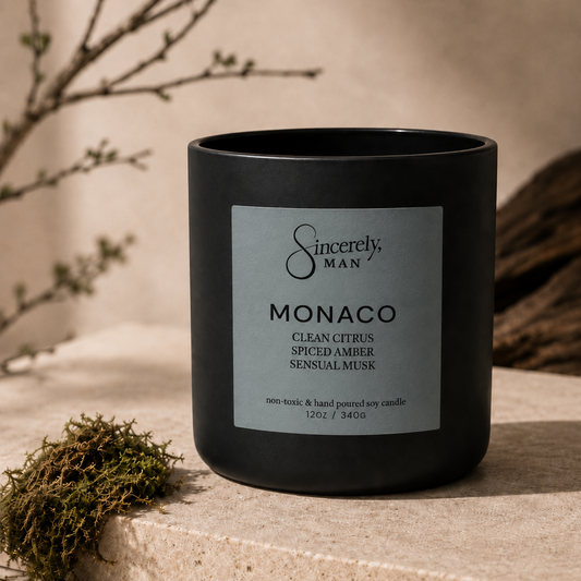 A matte black candle jar with a silver label reading 'Sincerely, MAN' and 'MONACO Clean Citrus Spiced Amber Sensual Musk', styled on a stone surface with moss and branches in a warm neutral setting.