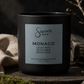 A matte black candle jar with a silver-gray label reading 'Sincerely, MAN' and 'MONACO', styled on a rustic wooden surface with moss, linen, and dried branches in a moody setting.
