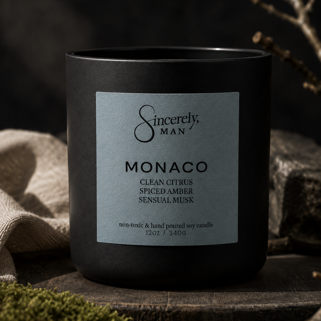 A matte black candle jar with a silver-gray label reading 'Sincerely, MAN' and 'MONACO', styled on a rustic wooden surface with moss, linen, and dried branches in a moody setting.