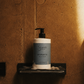 Bottle of Monaco body wash on a soap dish against a textured brown wall.