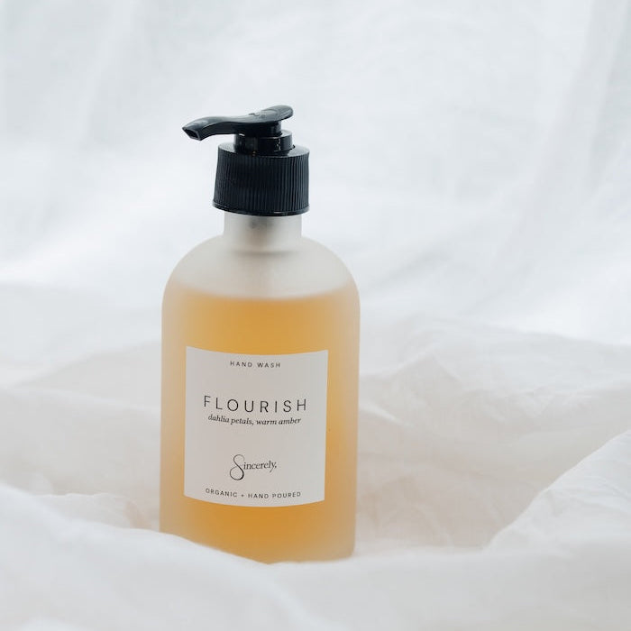 A frosted glass pump bottle of Flourish Hand Wash, filled with a light golden-yellow liquid, set against a draped white fabric background.