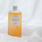 A clear glass bottle filled with amber-orange liquid, featuring a white label that reads 'FLOURISH' set against a soft white draped fabric background.