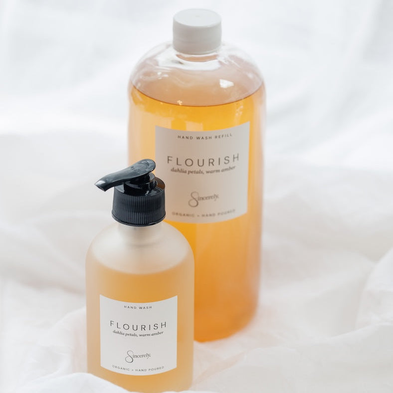 Two hand wash bottles labeled Flourish, one larger clear bottle with a white cap and one smaller frosted pump bottle, both filled with a golden-amber liquid, set against a draped white fabric background.