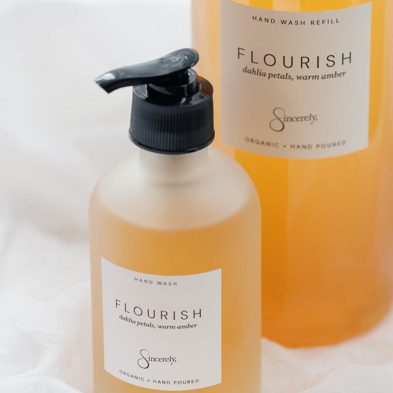 Two Sincerely brand hand wash refill bottles labeled 'Flourish' with a black pump dispenser in the foreground and a larger bottle in the background, set against a soft white fabric backdrop.