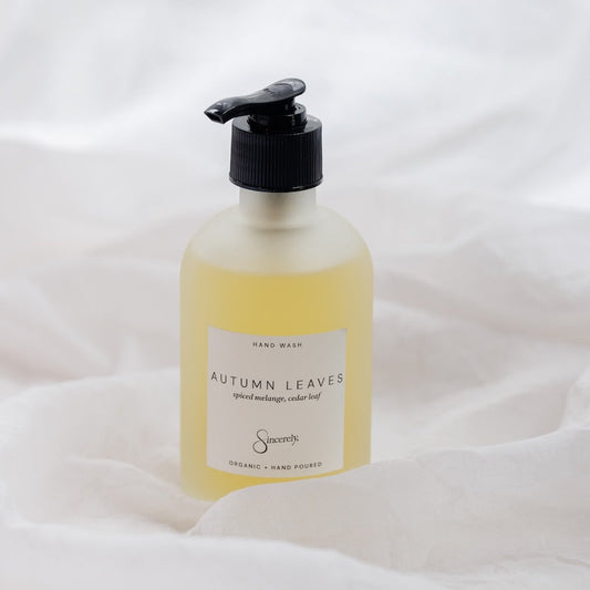 A frosted glass hand wash bottle with a black pump dispenser, filled with pale yellow liquid. The bottle features a white label with the text 'AUTUMN LEAVES' 
