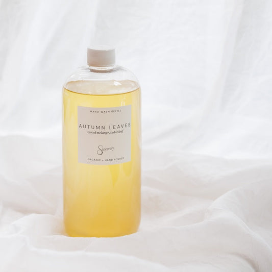 A clear bottle filled with pale golden-yellow liquid, standing upright on draped white fabric. The bottle has a white cap and a white label reading 'AUTUMN LEAVES' with smaller text above and below, and a small script logo near the bottom.