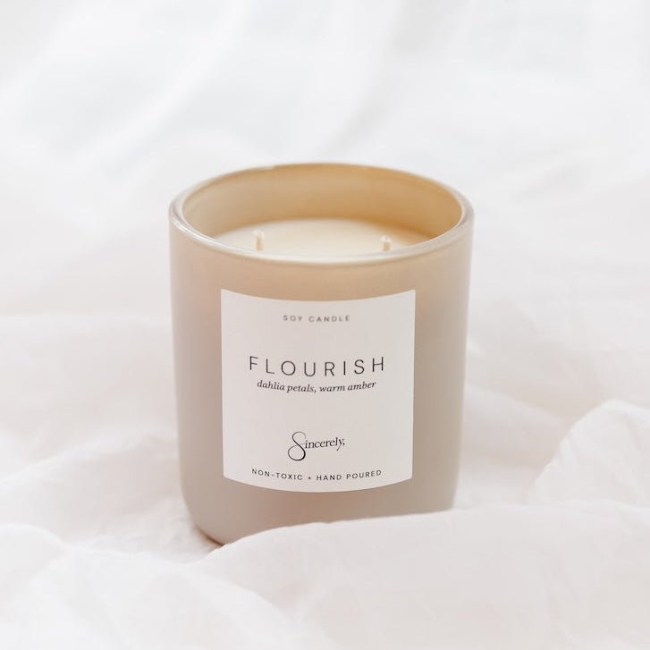 A soy candle in a matte taupe-beige glass jar with a white label reading 'FLOURISH', set against a soft white draped fabric background.