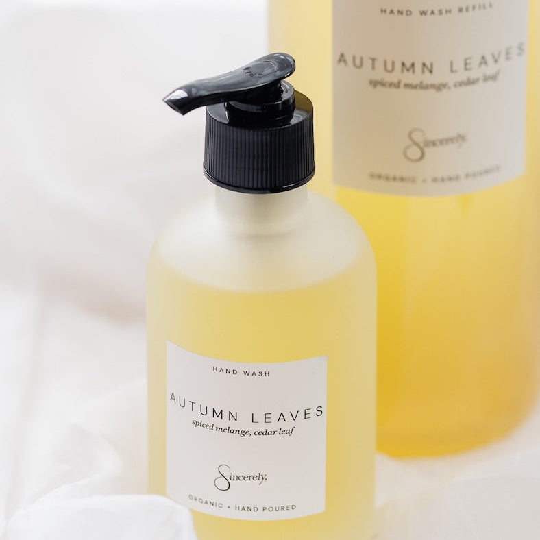 A frosted hand wash bottle with a black pump dispenser in the foreground and a larger bottle of the same product blurred in the background, both labeled Autumn Leaves set against a soft white fabric backdrop.