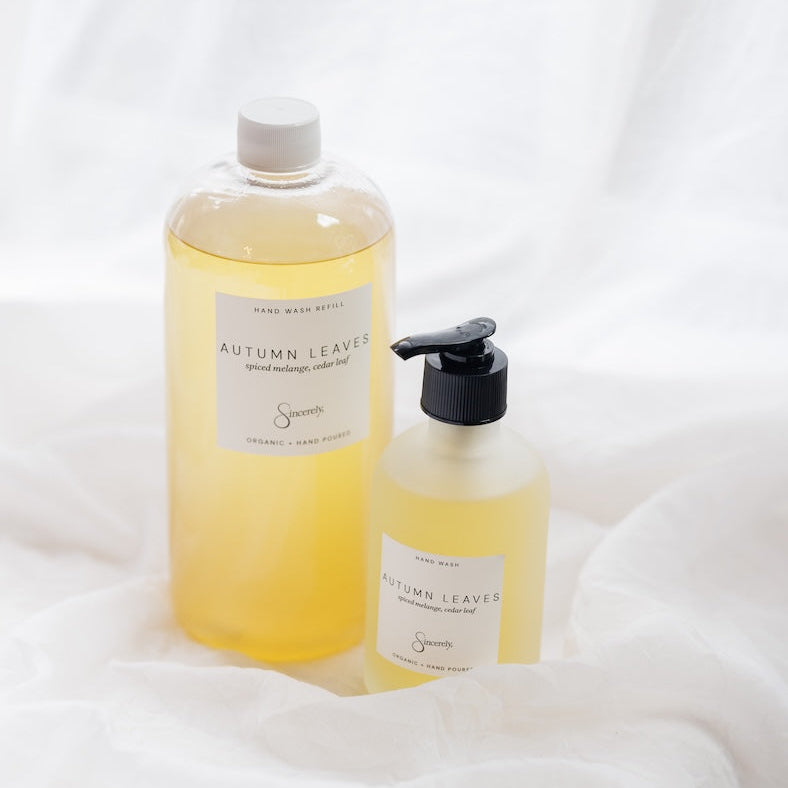 Two bottles of Autumn Leaves hand wash oil and hand wash liquid, filled with pale yellow liquid, displayed on a draped white fabric background.