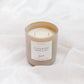 A two-wick soy candle in a frosted beige glass jar with a white label reading 'FLOURISH', set against a background of soft, draped white fabric.