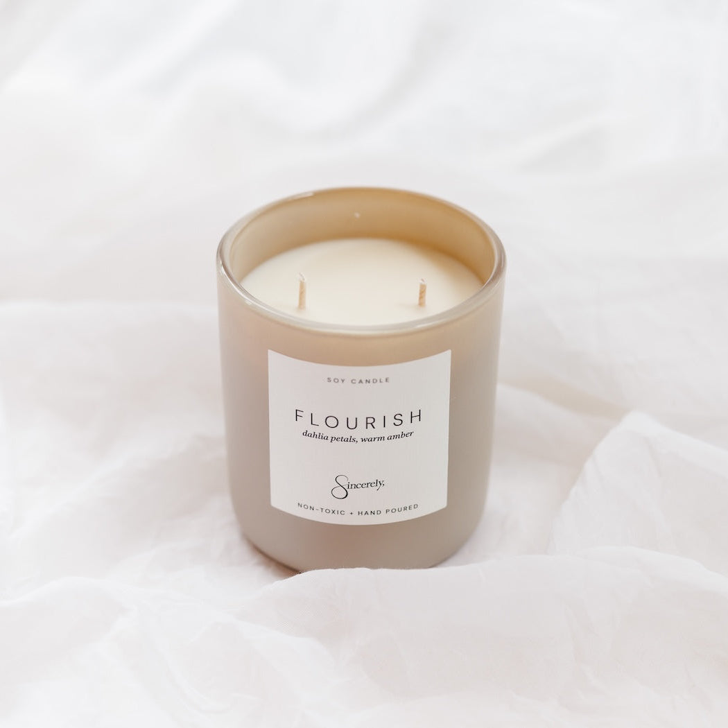 A two-wick soy candle in a frosted beige glass jar with a white label reading 'FLOURISH', set against a background of soft, draped white fabric.