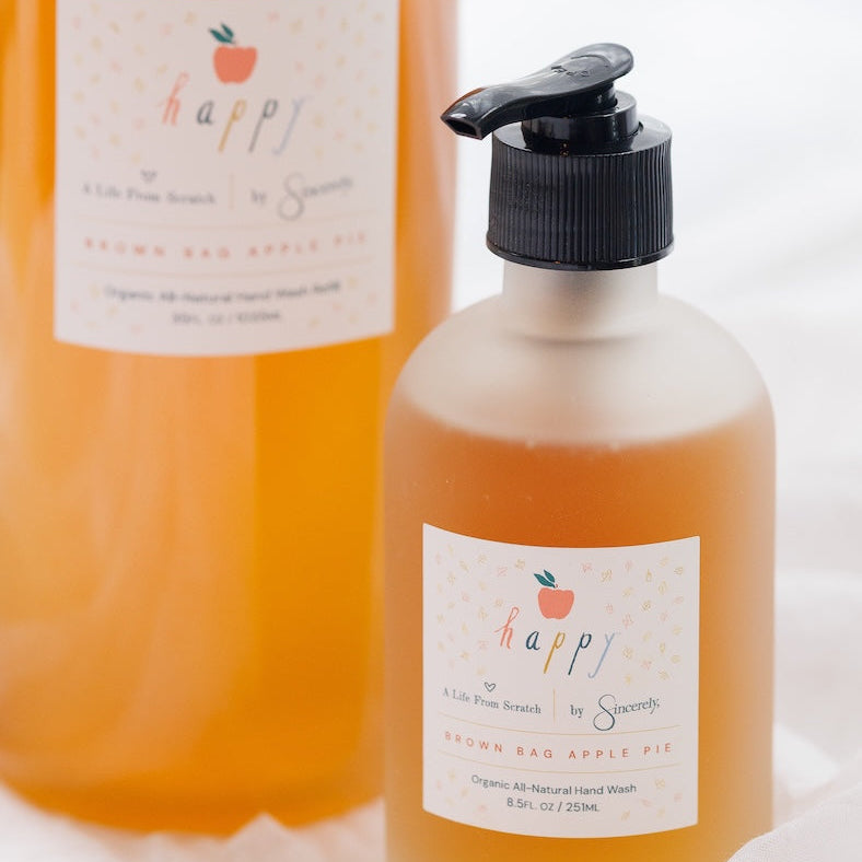 Two bottles of Happy Brown Bag Apple Pie scented hand wash, with one larger bottle in the background and a smaller pump bottle in the foreground on a white textured surface.
