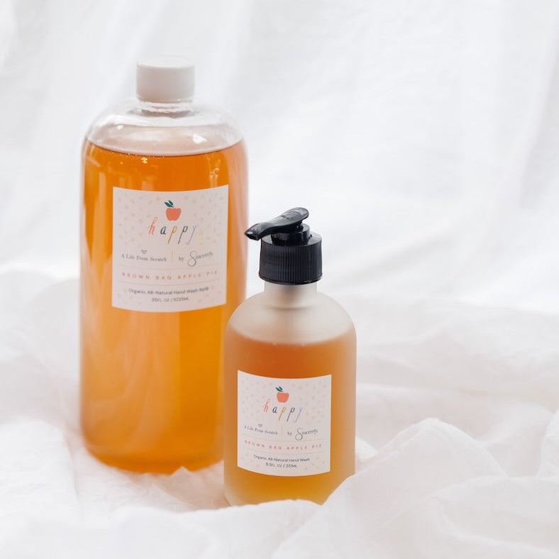 Two bottles of Happy Hand Washarranged on white draped fabric. The larger bottle on the left is filled with bright orange liquid and has a white cap.
