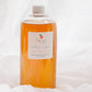 A clear plastic bottle filled with orange liquid, featuring a white cap and a white label with the brand name 'happy' and an apple graphic, set against a draped white fabric background.