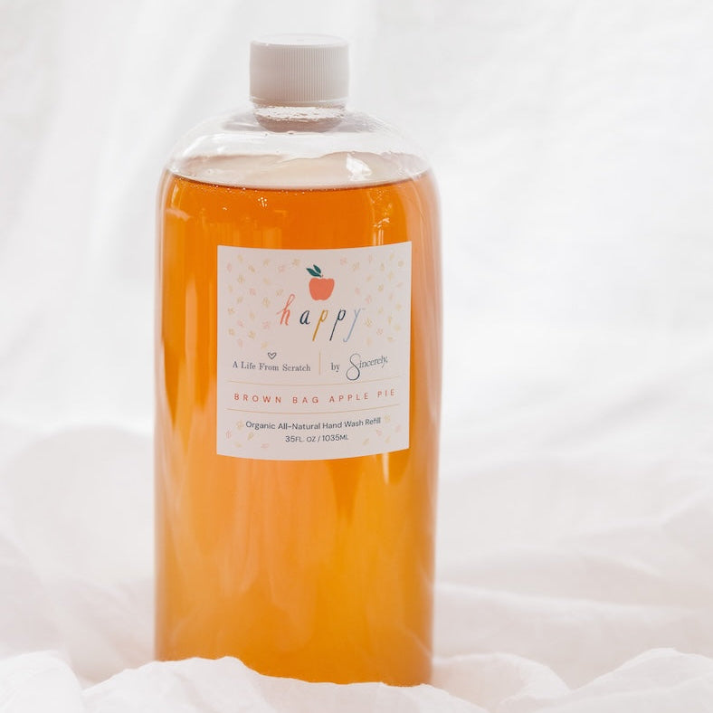 A clear plastic bottle filled with orange liquid, featuring a white cap and a white label with the brand name 'happy' and an apple graphic, set against a draped white fabric background.