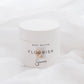 A white jar of Sincerely body butter labeled 'Flourish' resting on soft white draped fabric.